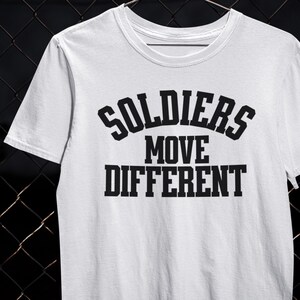 以下が含まれることがあります： 黒い文字で「SOLDIERS MOVE DIFFERENT」とプリントされた白いクルーネックTシャツ。テキストは3行に配置され、上部に「SOLDIERS」、中央に「MOVE」、下部に「DIFFERENT」と書かれています。シャツは半袖です。