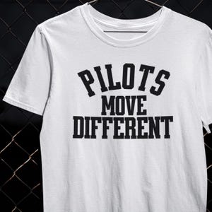 以下が含まれることがあります： 「PILOTS MOVE DIFFERENT」の文字が黒でプリントされた白いTシャツ。テキストは太字のサンセリフ体で、「PILOTS」、「MOVE」、「DIFFERENT」の3行に配置されています。Tシャツはクルーネックで半袖です。
