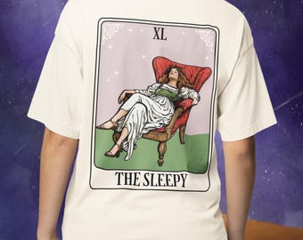 May include: Cream XL t-shirt with a tarot card design on the back. The card shows a person reclining in a red armchair, with the text "THE SLEEPY" below. The background is light pink with stars.