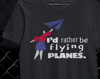 May include: Black t-shirt with a graphic of a person holding a blue paper airplane. The text "I'd rather be flying PLANES." is printed in white. The design features red, blue, and black colors.