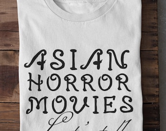 Może przedstawiać: Biała koszulka z czarnym napisem "ASIAN HORROR MOVIES Let's talk". Koszulka jest złożona i leży na drewnianej powierzchni.