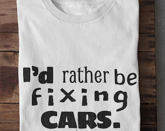 May include: White t-shirt with the text "I'd rather be fixing CARS" in black block letters. The shirt is folded on a wooden surface.