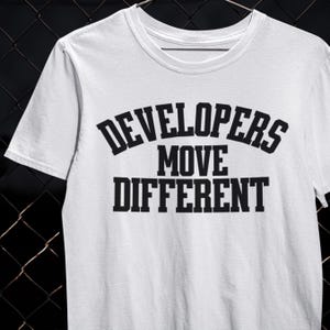 Op de afbeelding: Wit T-shirt met de zwarte tekst "DEVELOPERS MOVE DIFFERENT". De tekst is vetgedrukt, in hoofdletters en is in drie regels gerangschikt. Het T-shirt heeft een ronde hals en korte mouwen.