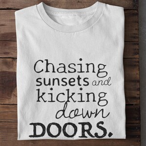 May include: White t-shirt with the text "Chasing sunsets and kicking down doors" in black lettering. The shirt is folded and placed on a wooden surface.
