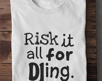 May include: White t-shirt with the phrase "Risk it all for Djing" printed in black, bold letters. The shirt is folded and rests on a wooden surface. A casual, everyday wear tee.