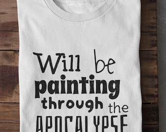 Puede incluir: Camiseta blanca doblada con el texto negro "Will be painting through the APOCALYPSE" en fuente negrita y estilizada. La camiseta está sobre una superficie de madera.