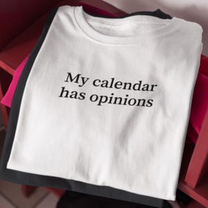 May include: A stack of t-shirts in white, black, and pink. The top white t-shirt has the phrase "My calendar has opinions" printed in black, centered on the front. The shirts are neatly folded and stacked on a red surface.