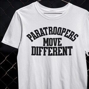 以下が含まれることがあります： 「PARATROOPERS MOVE DIFFERENT」のテキストが黒い太字でプリントされた白いTシャツ。テキストは3行に配置され、「PARATROOPERS」が「MOVE DIFFERENT」の上にアーチ状に配置されています。Tシャツはクルーネックで半袖です。