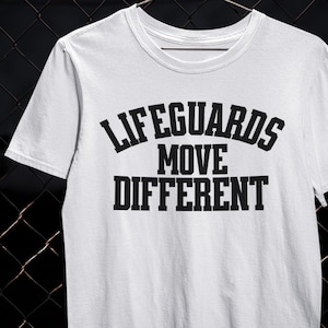 以下が含まれることがあります： 「LIFEGUARDS MOVE DIFFERENT」の文字が黒い太字でプリントされた白いTシャツ。Tシャツはクルーネックで半袖です。テキストは3行に配置されています。