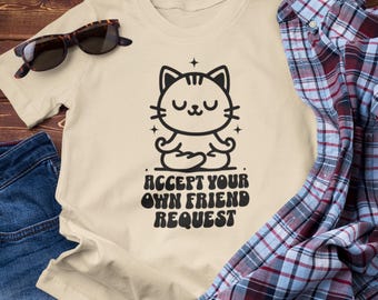 May include: A beige t-shirt featuring a black graphic of a meditating cat with the text "ACCEPT YOUR OWN FRIEND REQUEST". The shirt is paired with sunglasses, a blue denim garment, and a plaid shirt.