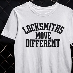 以下が含まれることがあります： 「LOCKSMITHS MOVE DIFFERENT」のテキストが黒い太字でプリントされた白いTシャツ。クルーネックと半袖のデザインです。テキストはシャツの前面中央に配置されています。