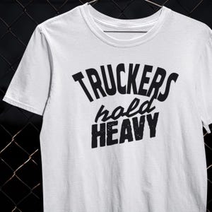 以下が含まれることがあります： 「TRUCKERS hold HEAVY」の黒い太字の文字がプリントされた白いクルーネックTシャツ。柔らかい素材でできており、カジュアルウェアに適しています。