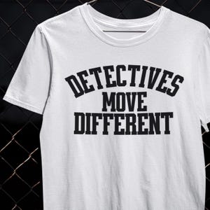 以下が含まれることがあります： 「DETECTIVES MOVE DIFFERENT」の文字が黒い太字でプリントされた白いTシャツ。クルーネックと半袖のデザインです。デザインは胸の中央に配置されています。