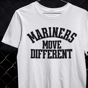 以下が含まれることがあります： 黒い太字のブロック文字で「MARINERS MOVE DIFFERENT」と書かれた白いTシャツ。Tシャツはクルーネックと半袖です。シャツは柔らかく快適な素材で作られています。
