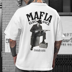 May include: White t-shirt featuring a black and white image of a man in a suit with a gun, the words "MAFIA ELEGANCE" above, and "Criminally stylish." below the image.