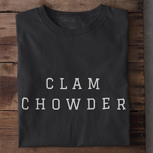 Puede incluir: Camiseta negra con las palabras "CLAM CHOWDER" impresas en letras blancas. La camiseta está doblada y sobre una superficie de madera.