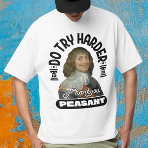Può includere: T-shirt bianca con una grafica raffigurante un uomo e il testo "DO TRY HARDER" e "Thankyou PEASANT" in un ovale nero. Il disegno è centrato sulla maglietta.