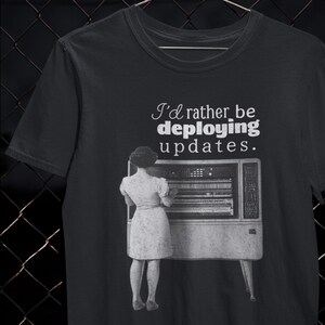 May include: Black t-shirt with the text "I'd rather be deploying updates." The shirt features a vintage-style image of a person standing in front of a retro computer. The design is in white and the shirt is a dark colour.