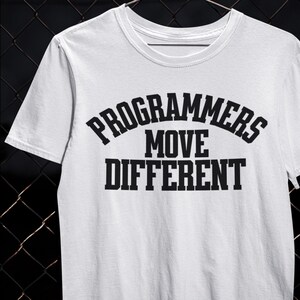 Pode incluir: Camiseta branca com o texto preto "PROGRAMMERS MOVE DIFFERENT" em letras maiúsculas e em negrito. A camiseta tem gola redonda e mangas curtas. O design está centralizado na frente.
