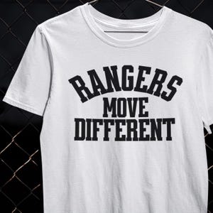以下が含まれることがあります： 「RANGERS MOVE DIFFERENT」のテキストが黒色の太字でプリントされた白いTシャツ。クルーネックと半袖のデザインです。デザインは胸の中央に配置されています。