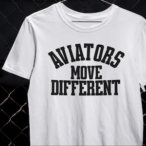 以下が含まれることがあります： 「AVIATORS MOVE DIFFERENT」のテキストが黒い太字でプリントされた白いTシャツ。クルーネックと半袖のデザイン。テキストは3行に配置され、上部に「AVIATORS」とアーチ状に配置されています。