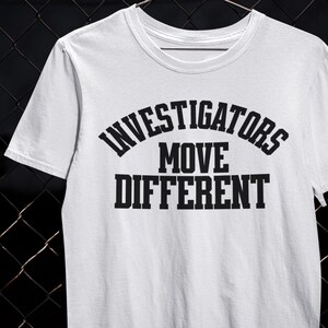 以下が含まれることがあります： 「INVESTIGATORS MOVE DIFFERENT」の黒い文字がプリントされた白いTシャツ。 「INVESTIGATORS」の文字は「MOVE DIFFERENT」の上にアーチ状に配置されています。 このシャツはクルーネックで半袖です。