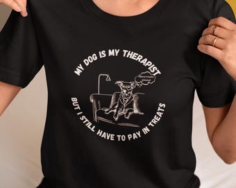 Könnte beinhalten: Schwarzes T-Shirt mit einem weißen Grafikdesign, auf dem "Mein Hund ist mein Therapeut, aber ich muss trotzdem in Leckerlis bezahlen" steht. Die Grafik zeigt auch einen Cartoon-Hund, der auf einem Sofa sitzt, mit einer Gedankenblase, in der "Mehr Leckerlis bitte!" steht.