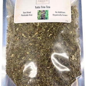 May include: A clear plastic bag filled with dried green herbs. The label reads "Tulsi Trio Tea," with text indicating it's sun-dried, pesticide-free, and organically grown. The bag contains 2 oz of tea.