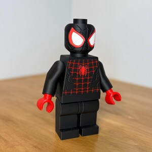 May include: A black Lego-style figure of a superhero with red and white eyes, red hands, and a red spiderweb design on the chest. The figure is standing on a wooden surface.