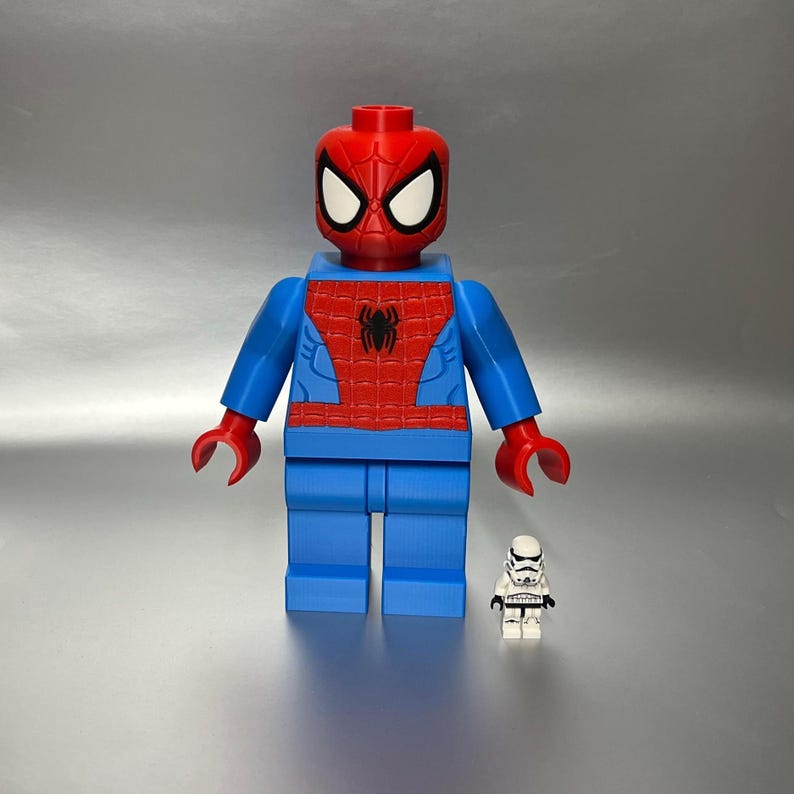 May include: A large, red, blue, and black Spider-Man figure, resembling a Lego toy, stands next to a smaller white Stormtrooper figure. The Spider-Man figure has a red head and torso with a black spider emblem.