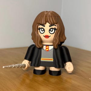May include: A 3D-printed figurine of a character with brown hair, red lips, and a gray sweater with a red and yellow tie. The figure wears a black robe and holds a silver wand. The figurine is on a wooden surface.