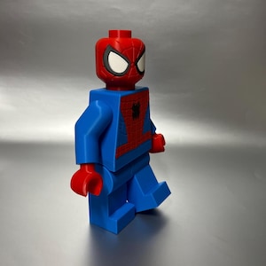 May include: A large, blocky figure of Spider-Man in a walking pose. The figure is primarily blue with red accents on the head, chest, and hands. The head features the iconic Spider-Man mask with white eyes and a red spider emblem.