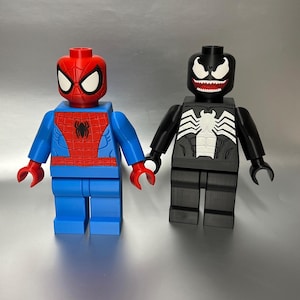 May include: Two large, blocky figures resembling Lego minifigures. One is Spider-Man, with a red head and torso, blue arms and legs. The other is Venom, black with a white spider emblem and teeth. Both figures are holding hands.