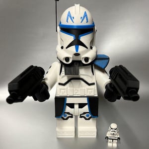 May include: A large white and blue Clone Trooper figure with two black blasters, and a smaller matching figure. The large figure has a blue and black design on the helmet and armour. The figures are on a grey background.
