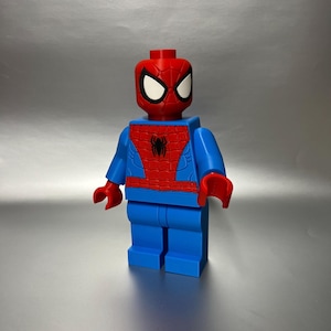 May include: A large, blue and red Spider-Man figure, resembling a Lego toy. The figure has a red head with white eye details, a red torso with a black spider emblem, and blue arms and legs. The hands are red.