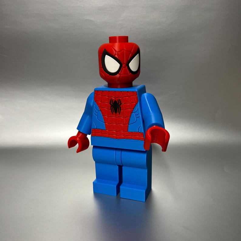 May include: A large, blue and red Spider-Man figure, resembling a Lego toy. The figure has a red head with white eye details, a red torso with a black spider emblem, and blue arms and legs. The hands are red.