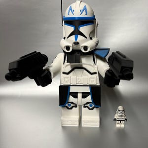 May include: A large white and blue Lego-style figure of a Star Wars clone trooper, holding two black blasters. A smaller clone trooper figure is next to it. The figures are on a silver background.