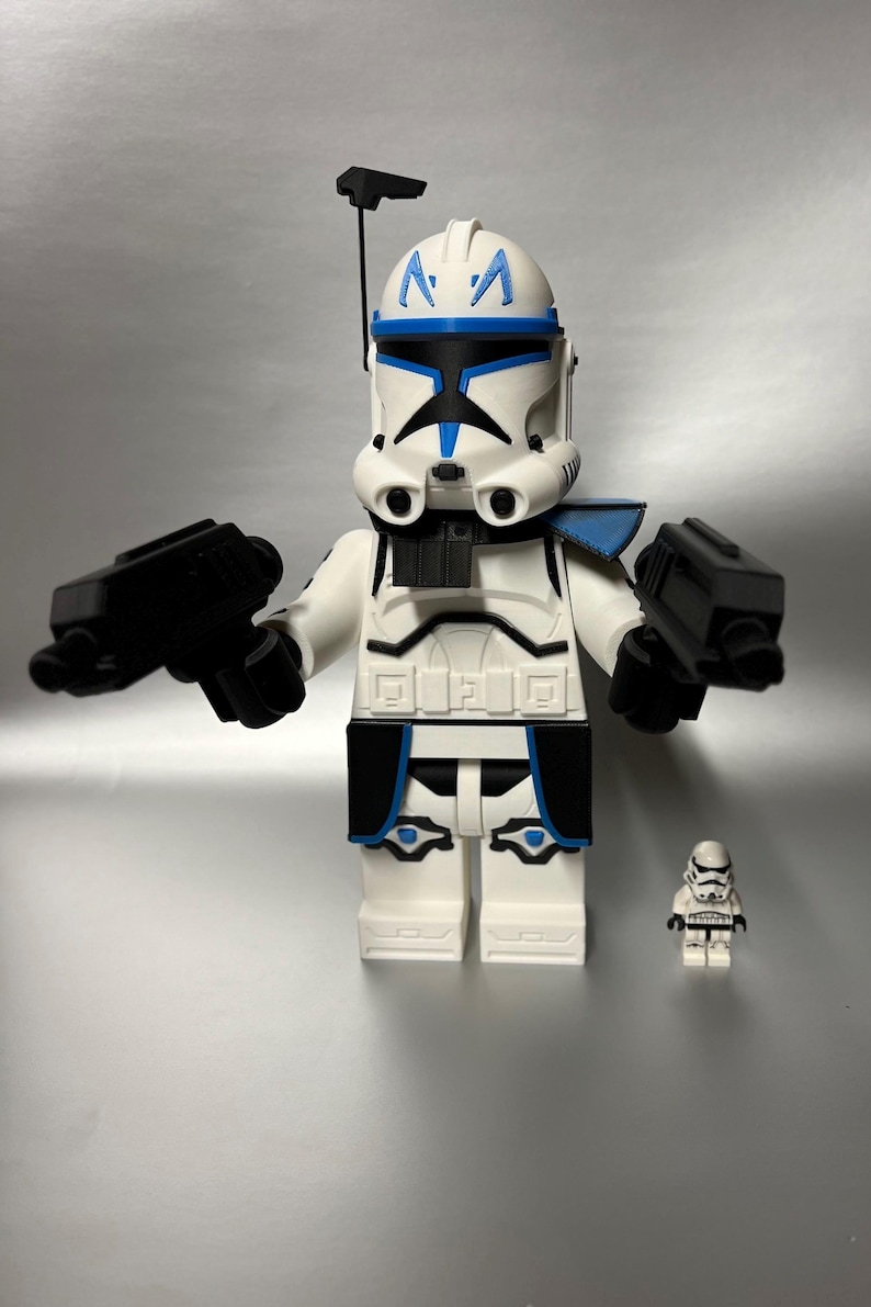 May include: A large white and blue Lego-style figure of a Star Wars clone trooper, holding two black blasters. A smaller clone trooper figure is next to it. The figures are on a silver background.