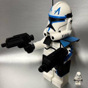 May include: A large white and blue clone trooper action figure, holding two black blasters. The figure has a detailed helmet with blue markings and a small antenna. A smaller clone trooper figure is also present.