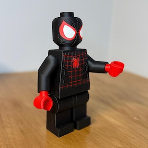 May include: A black and red Spider-Man figure, styled like a Lego minifigure. The figure features a black body with red detailing, including the eyes, hands, and a spider emblem. The figure is posed with one arm outstretched.