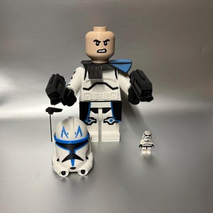 May include: A large white and blue Lego figure with a matching helmet and two black blasters. A smaller Lego figure and helmet are also present. The large figure has a neutral skin tone and is wearing a white and black uniform.