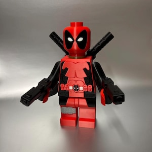 May include: A red and black Deadpool figure with two black swords on its back and two black guns. The figure has a red head with white eyes and a black mask. The figure is a toy.