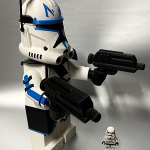 May include: A large white and blue clone trooper action figure holding two black blasters, with a smaller clone trooper figure in the background. The larger figure has a detailed helmet with blue markings and a black antenna.