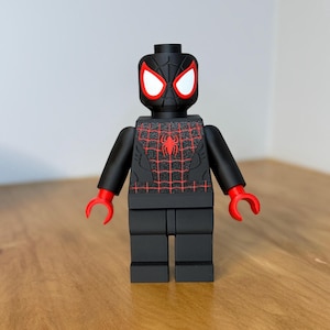 May include: A black Lego-style Spider-Man figure with red detailing. The figure has a red spider emblem on the chest and red web detailing. The eyes are white, and the hands are red. The figure is on a wooden surface.