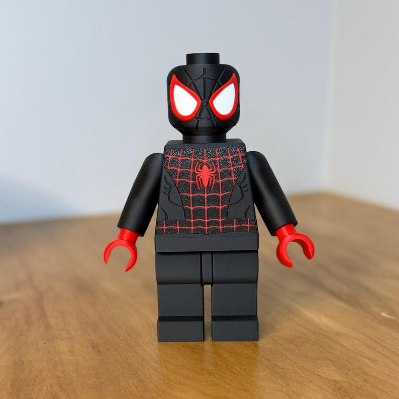 May include: A black Lego-style Spider-Man figure with red detailing. The figure has a red spider emblem on the chest and red web detailing. The eyes are white, and the hands are red. The figure is on a wooden surface.