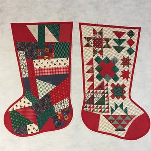 May include: Two Christmas stockings with red trim. One stocking features a patchwork design with various patterns in red, green, and white. The other stocking has a cream background with red and green geometric shapes and a basket design.