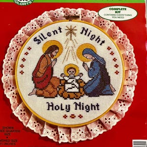 May include: A stamped cross stitch kit featuring a Christmas scene with the text "Silent Night Holy Night." The kit includes a wooden hoop, fabric, embroidery floss, and needle. The finished size is 10.5 inches.