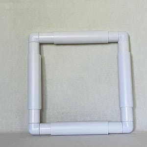 May include: A white, square embroidery hoop, assembled from plastic tubes and right-angle connectors. The frame is designed to hold fabric taut for needlework projects. The clean, simple design is suitable for various crafting applications.