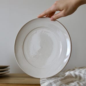 May include: A white ceramic dinner plate with a slightly raised rim. The plate has a subtle, textured surface.