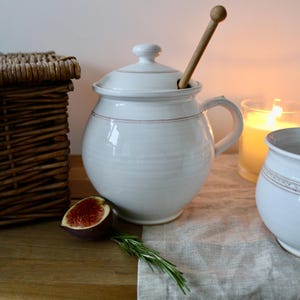 Może przedstawiać: Biały ceramiczny słoik na miód z pokrywką i uchwytem, ​​z drewnianym nabierakiem do miodu. Widoczny jest również wiklinowy kosz i mała miska. Figa i gałązka rozmarynu leżą na drewnianej powierzchni.